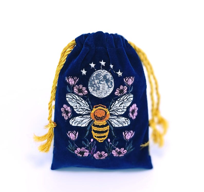 May include: A deep blue velvet drawstring pouch featuring an embroidered design of a central bee with yellow and black stripes, white wings, a full moon above, small stars, and pink flowers surrounding the bee on the front surface.