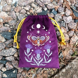 May include: A purple drawstring bag with a celestial moth and thistle embroidery design. The moth is orange and brown, with white stars and crescent moons above it. The bag has yellow drawstrings.