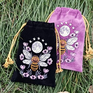 May include: Two velvet drawstring bags, one black and one purple, embroidered with a bee, moon, stars, and flowers. The black bag has a gold drawstring, and the purple bag has a gold drawstring. The bags are on a bed of green grass.
