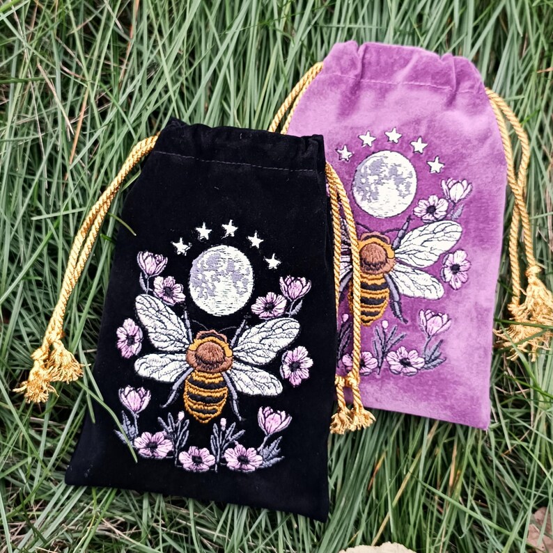 May include: Two velvet drawstring bags, one black and one purple, embroidered with a bee, moon, stars, and flowers. The black bag has a gold drawstring, and the purple bag has a gold drawstring. The bags are on a bed of green grass.