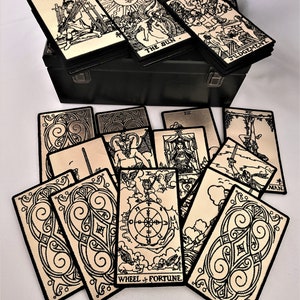 May include: A set of tarot cards with black and white illustrations on a gold background. The cards are arranged in a fan shape and are surrounded by a black border. The cards are in a metal box with a black finish. The cards feature the names of the cards, such as "The Sun", "Judgement", and "Wheel of Fortune".
