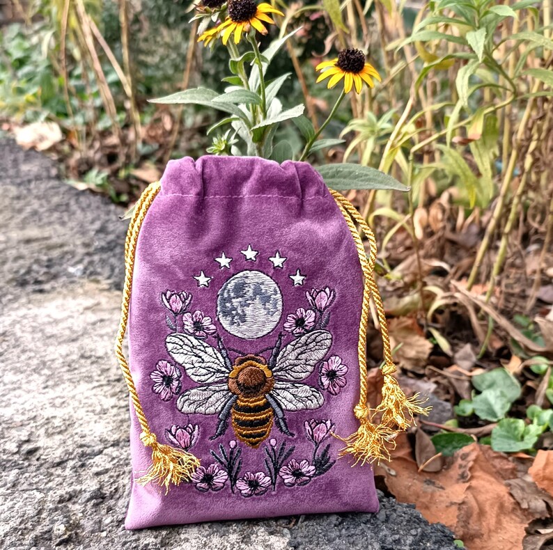 May include: A purple velvet drawstring bag with an embroidered bee design. The bee is centered below a full moon and surrounded by flowers and stars. The bag has gold-colored drawstrings with tassels.