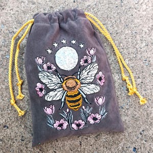 May include: A dark gray velvet drawstring bag with an embroidered design. The design features a large bee with white wings and a yellow and black striped body, surrounded by pink flowers, a moon, and stars. The drawstring is gold.