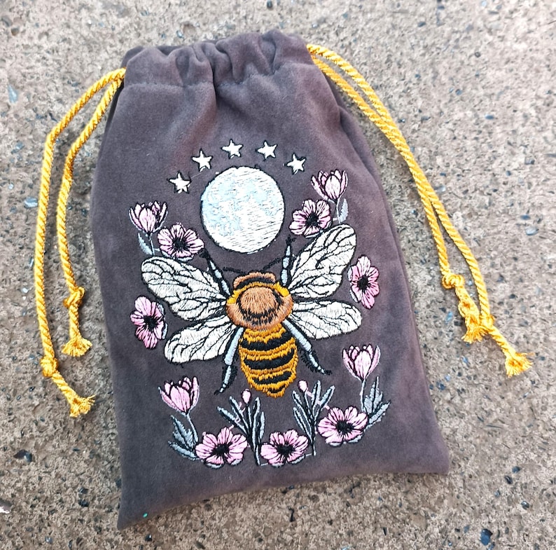 May include: A dark gray velvet drawstring bag with an embroidered design. The design features a large bee with white wings and a yellow and black striped body, surrounded by pink flowers, a moon, and stars. The drawstring is gold.