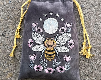 May include: A gray drawstring pouch featuring an embroidered design of a bee centered below a white moon and stars, surrounded by pink flowers with green stems. The bag has two yellow braided cords that pull to close the top opening.