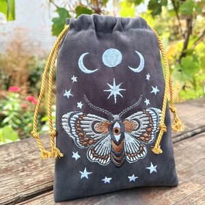 May include: Dark grey velvet drawstring bag with gold cord. The bag features an embroidered design of a moth with a central eye, surrounded by stars, crescent moons, and a full moon. The moth has brown, grey, and white details.