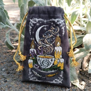 May include: A dark grey velvet drawstring bag with an embroidered design featuring a crescent moon, teacup, candles, and herbs. The bag has a gold drawstring closure and tassel ends. The embroidery is in white, gold, and purple.