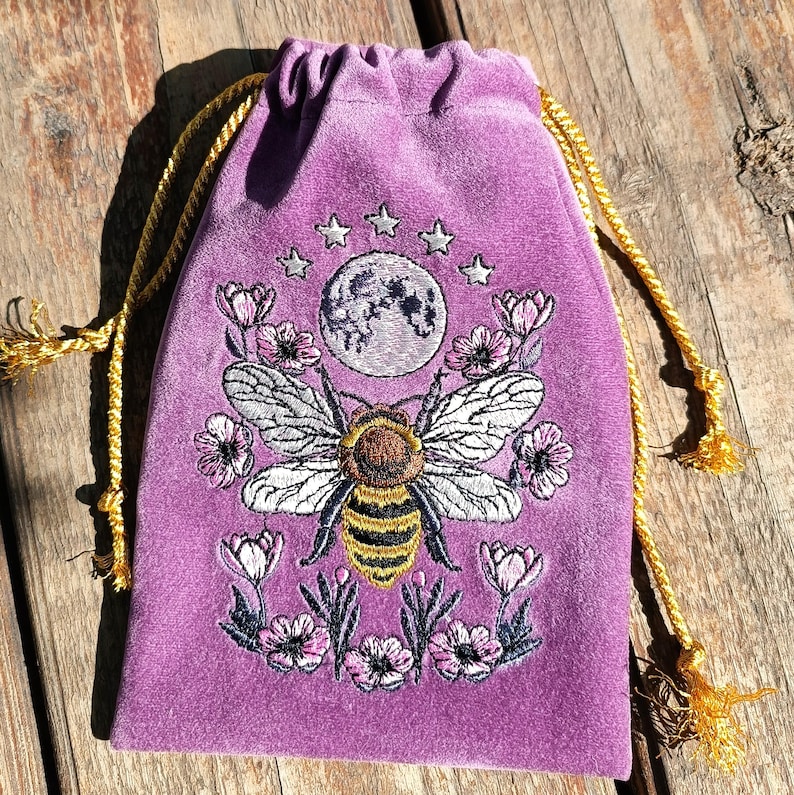 May include: A purple velvet drawstring bag with an embroidered design. The design features a large bee with detailed wings, surrounded by flowers, a full moon, and stars. The drawstring is gold.