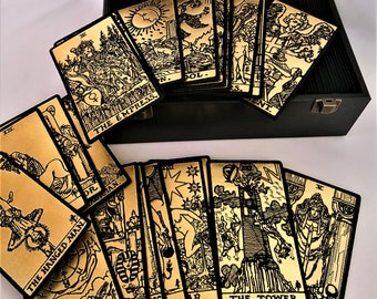 May include: A set of gold tarot cards with black illustrations and text. The cards are laid out in a fan shape on a black surface. The cards include "The Hanged Man", "The Empress", "Wheel & Fortune", "The Tower", and "The Chariot".