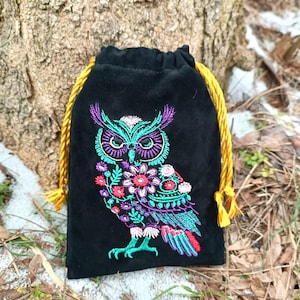 May include: Black drawstring bag with an embroidered owl design. The owl is depicted with teal, purple, pink, and red floral details. The bag has a gold drawstring closure and is made of a soft, velvety material.