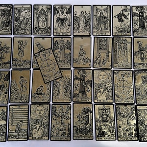 May include: A full deck of tarot cards with black and white illustrations on a gold background. The cards are arranged in rows and columns, with the backs of the cards facing up. The cards are numbered from 1 to 22, and each card has a different illustration.