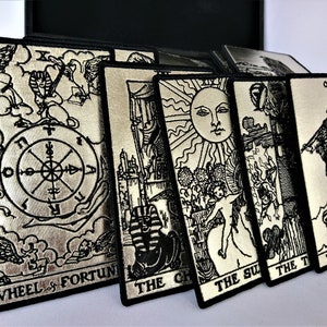 May include: A set of silver tarot card patches with black embroidery. The patches feature the Wheel of Fortune, The Chariot, The Sun, The Tower, and The Fool cards.