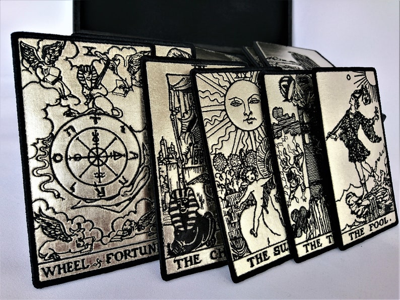 May include: A set of silver tarot card patches with black embroidery. The patches feature the Wheel of Fortune, The Chariot, The Sun, The Tower, and The Fool cards.