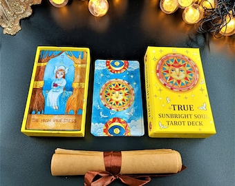 May include: A yellow box with the text "TRUE SUNBRIGHT SOUL TAROT DECK" on the front. The box contains a tarot deck with colourful illustrations of the sun and other celestial bodies. The box is sitting on a black surface with a roll of brown paper and a string of lights in the background.