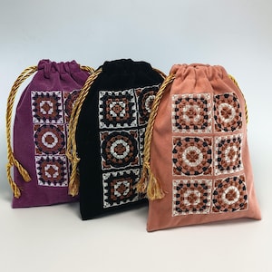 Puede incluir: Tres bolsas con cordón de terciopelo en color morado, negro y rosa empolvado. Cada bolsa presenta un panel frontal con seis motivos cuadrados bordados en blanco, marrón y negro. Las bolsas se cierran con cordones trenzados dorados.