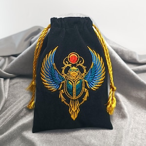 Puede incluir: Una bolsa negra con cordón ajustable que presenta un diseño bordado de un escarabajo alado. El escarabajo está realizado con hilo dorado, azul y turquesa, rematado con un círculo rojo. La bolsa se cierra con dos cordones dorados trenzados.