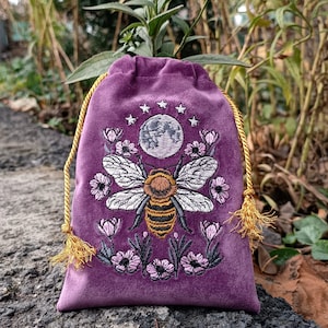 May include: A purple velvet drawstring bag with an embroidered design featuring a large bee, a full moon, stars, and flowers. The bag has a gold drawstring closure with tassels. The embroidery is in shades of white, brown, black, and pink.