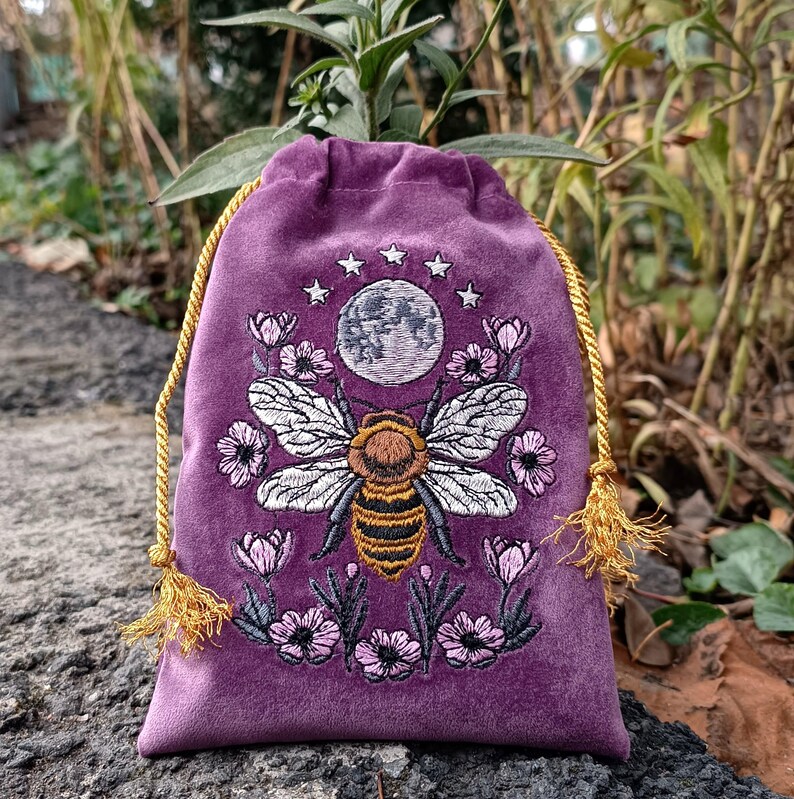 May include: A purple velvet drawstring bag with an embroidered design featuring a large bee, a full moon, stars, and flowers. The bag has a gold drawstring closure with tassels. The embroidery is in shades of white, brown, black, and pink.