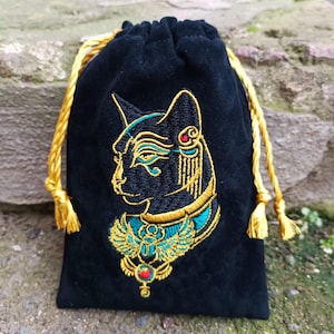 May include: A black velvet drawstring pouch featuring an embroidered design of an Egyptian cat deity in profile. The embroidery uses gold, teal, and red thread to depict the cat wearing an ornate collar with a winged scarab and a central red gem.