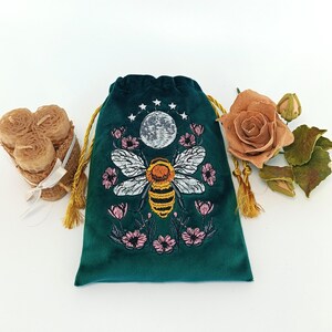 May include: A dark green velvet drawstring pouch featuring an embroidered design of a central bee, a moon, and surrounding pink flowers. The pouch has gold-colored drawstrings and is positioned between a bundle of beeswax candles and a dried rose.