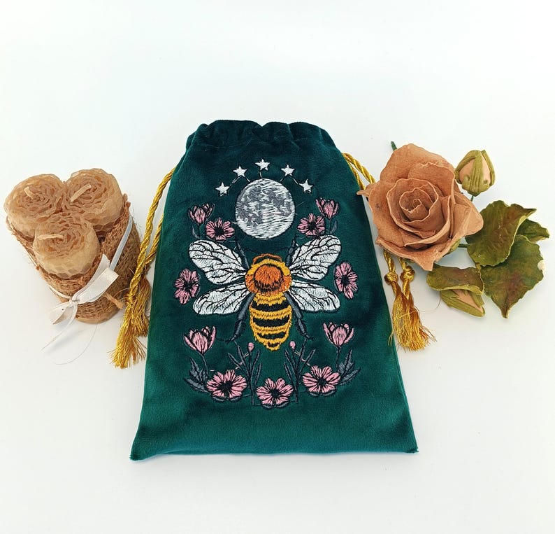 May include: A dark green velvet drawstring pouch featuring an embroidered design of a central bee, a moon, and surrounding pink flowers. The pouch has gold-colored drawstrings and is positioned between a bundle of beeswax candles and a dried rose.