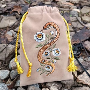 May include: A tan drawstring bag with a detailed embroidered design. The design features a brown and orange snake intertwined with white and yellow flowers. The bag has a yellow drawstring closure.