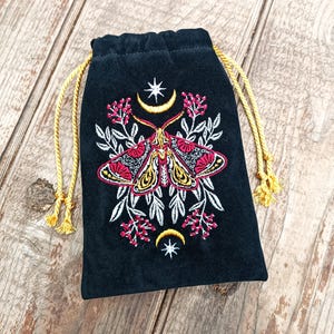 Puede incluir: Bolsa negra de terciopelo con cordón y diseño de polilla bordada. La polilla tiene detalles rojos, amarillos y negros, rodeada de hojas blancas y bayas rosas. Cierre dorado con cordón.