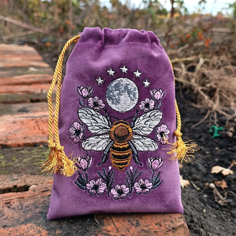 May include: A purple velvet drawstring bag with an embroidered design. The design features a large bee with detailed wings, surrounded by flowers, a full moon, and stars. The drawstring is gold, and the bag is on a brick surface.