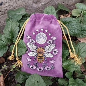 May include: A purple velvet drawstring bag with a detailed embroidered design. The design features a large bee with a full moon, stars, and floral accents. The bag has gold-colored drawstrings and tassels, set against a backdrop of green foliage.