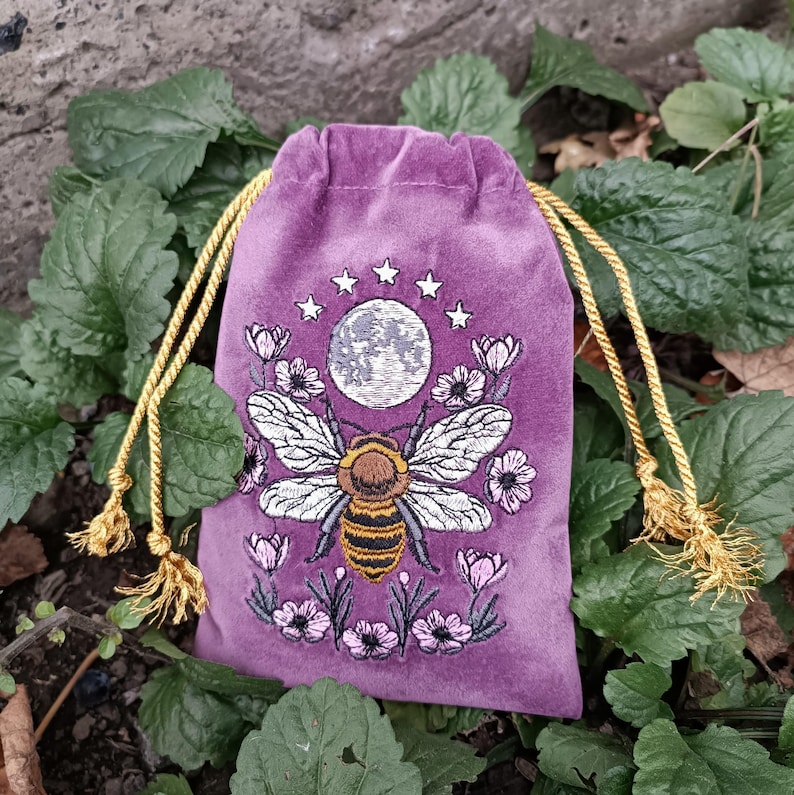May include: A purple velvet drawstring bag with a detailed embroidered design. The design features a large bee with a full moon, stars, and floral accents. The bag has gold-colored drawstrings and tassels, set against a backdrop of green foliage.