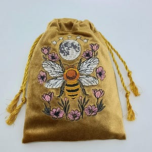 May include: A gold velvet drawstring pouch featuring an embroidered design of a central bee with white wings, surrounded by pink flowers and a grey moon with small stars above. The bag includes matching gold twisted cord drawstrings with tassels.
