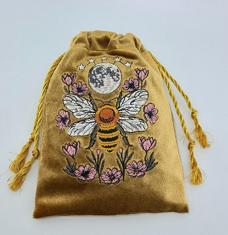 May include: A gold velvet drawstring pouch featuring an embroidered design of a central bee with white wings, surrounded by pink flowers and a grey moon with small stars above. The bag includes matching gold twisted cord drawstrings with tassels.