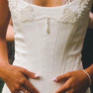 May include: A white corset top featuring a sweetheart neckline with delicate lace trim, intricate white floral embroidery, and small pearl bead embellishments. A single teardrop-shaped pearl pendant hangs from the center of the bodice.