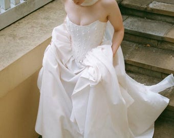 May include: A white strapless gown featuring a structured corset bodice with subtle beadwork and a voluminous, floor-length skirt with deep folds. The garment is paired with yellow pointed-toe heels and a matching white sheer scarf draped around the neck.