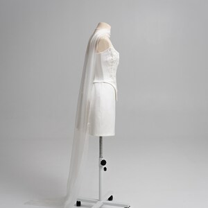 Puede incluir: Un vestido corto blanco sin tirantes con un corpi&ntilde;o estructurado que presenta intrincados bordados florales y un dobladillo en punta. Un velo blanco largo, transparente y fluido est&aacute; sujeto al escote y cae hasta el suelo.