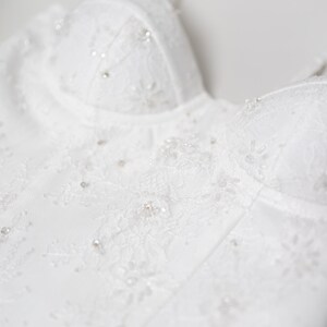 May include: Close-up of a white wedding dress bodice. The bodice features intricate lace detailing and is embellished with small, sparkling beads. The design includes a sweetheart neckline and delicate straps, creating an elegant and romantic look.