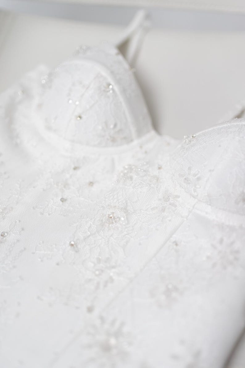 May include: Close-up of a white wedding dress bodice. The bodice features intricate lace detailing and is embellished with small, sparkling beads. The design includes a sweetheart neckline and delicate straps, creating an elegant and romantic look.
