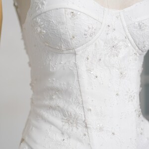 May include: A white wedding dress bodice with a sweetheart neckline. The dress features intricate lace detailing and is embellished with small, sparkling beads. The bodice is structured and fitted, creating a classic and elegant silhouette.