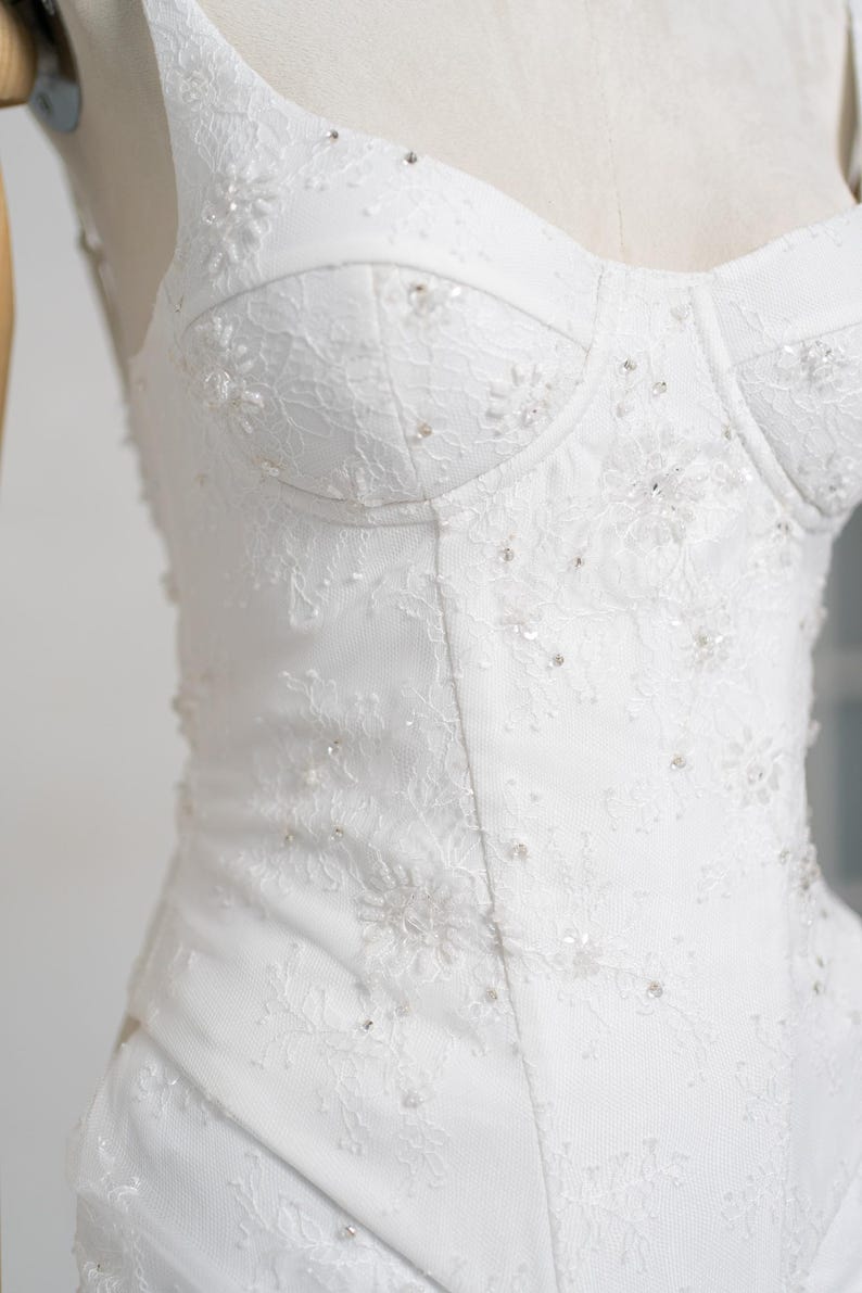 May include: A white wedding dress bodice with a sweetheart neckline. The dress features intricate lace detailing and is embellished with small, sparkling beads. The bodice is structured and fitted, creating a classic and elegant silhouette.
