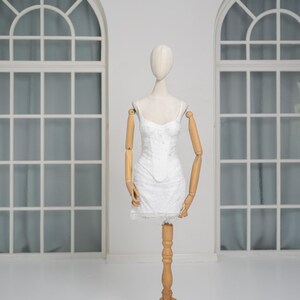 May include: A white, floral-patterned mini dress on a wooden mannequin. The dress features spaghetti straps and a lace hem. The mannequin has wooden arms and is displayed in a bright, white room with arched windows.