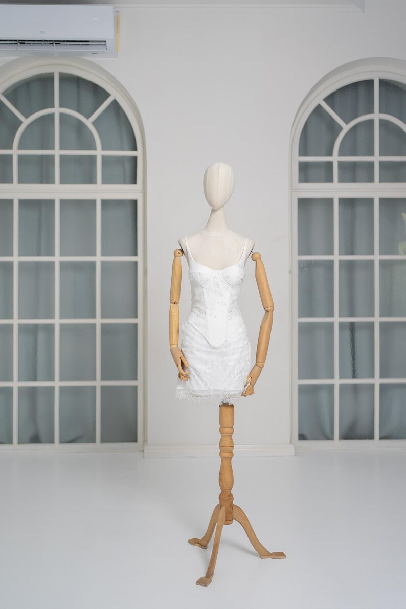 May include: A white, floral-patterned mini dress on a wooden mannequin. The dress features spaghetti straps and a lace hem. The mannequin has wooden arms and is displayed in a bright, white room with arched windows.