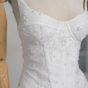 May include: White wedding dress with a fitted bodice and spaghetti straps. The bodice is embellished with delicate lace and sparkling bead accents. The dress is displayed on a mannequin, showcasing its elegant design and intricate details.