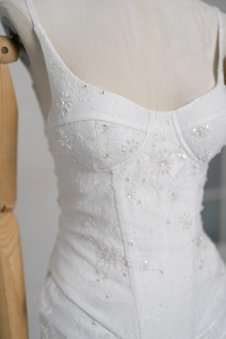 May include: White wedding dress with a fitted bodice and spaghetti straps. The bodice is embellished with delicate lace and sparkling bead accents. The dress is displayed on a mannequin, showcasing its elegant design and intricate details.