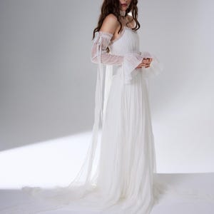 May include: A floor-length white dress featuring a pleated bodice, a straight neckline, and long, sheer, detachable sleeves with ruffled cuffs and long ribbon ties. The skirt is made of layered, flowing fabric that extends into a train.