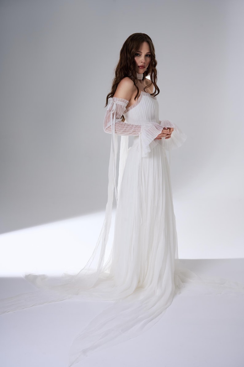 May include: A floor-length white dress featuring a pleated bodice, a straight neckline, and long, sheer, detachable sleeves with ruffled cuffs and long ribbon ties. The skirt is made of layered, flowing fabric that extends into a train.