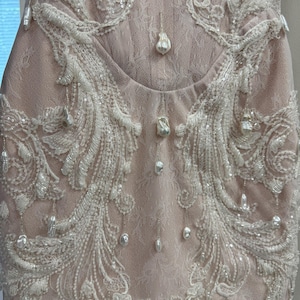 Pu&ograve; includere: Un abito senza maniche color rosa pallido con intricati strati di pizzo bianco e ricami elaborati. Il capo &egrave; decorato con numerose perle barocche bianche pendenti e piccole paillettes trasparenti che creano un aspetto stratificato.