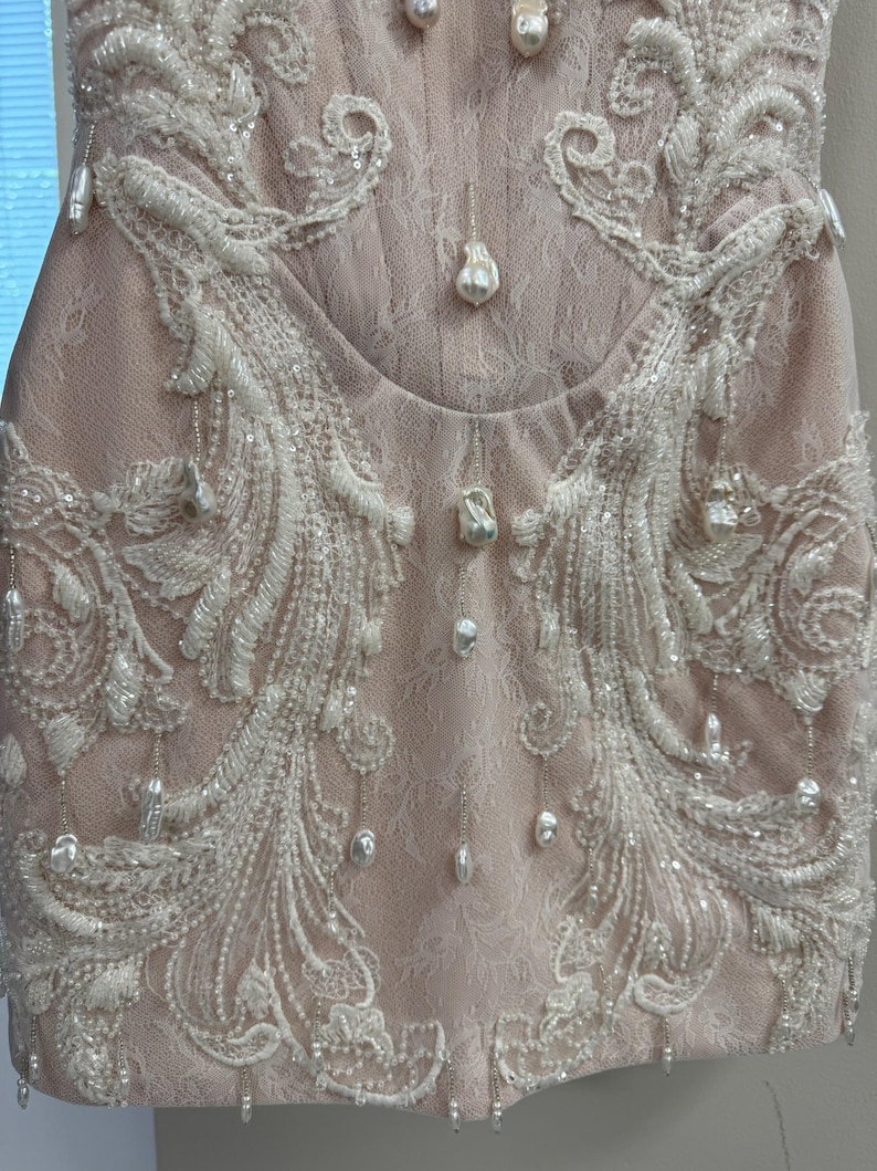 Pu&ograve; includere: Un abito senza maniche color rosa pallido con intricati strati di pizzo bianco e ricami elaborati. Il capo &egrave; decorato con numerose perle barocche bianche pendenti e piccole paillettes trasparenti che creano un aspetto stratificato.