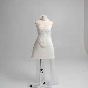 Puede incluir: Un vestido corto blanco sin tirantes con un corpi&ntilde;o estructurado que presenta intrincados bordados florales y pedrer&iacute;a. La prenda incluye un accesorio transparente y fluido tipo bufanda drapeado alrededor del cuello y que cae por la espalda.