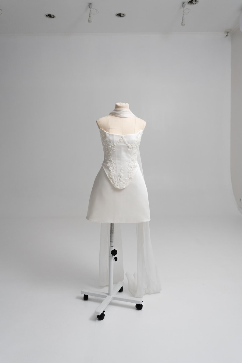 Puede incluir: Un vestido corto blanco sin tirantes con un corpi&ntilde;o estructurado que presenta intrincados bordados florales y pedrer&iacute;a. La prenda incluye un accesorio transparente y fluido tipo bufanda drapeado alrededor del cuello y que cae por la espalda.