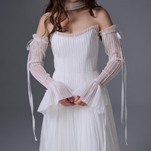 May include: A white floor-length dress featuring a pleated bodice and skirt, paired with matching sheer, pleated, off-the-shoulder long sleeves that have tie-up details at the upper arms and flared cuffs, along with a coordinating sheer neck scarf.